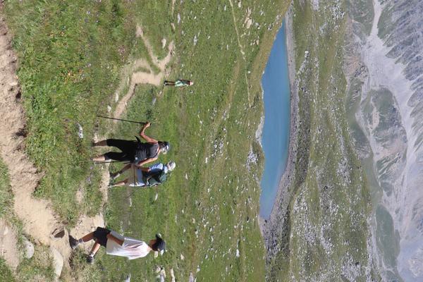 Four individuals are seen hiking along a winding dirt path on a grassy mountain slope, with one person using hiking poles